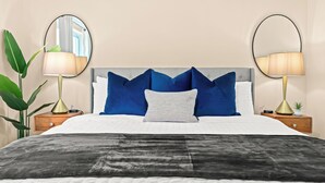 Un lit bien fait, recouvert d’une couverture grise et noire, deux tables de chevet avec des lampes, et un grand miroir de chaque côté.