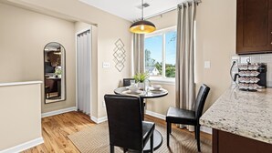 Un coin repas avec une table ronde, des chaises noires, un luminaire suspendu et une vue sur l’extérieur par une fenêtre ornée de rideaux.
