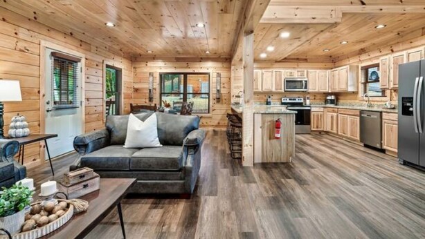A cabin interior with a living area featuring a sofa and coffee table, wooden floors, and a kitchen with modern appliances. - NEW BUILD SEPT 2023 4 KINGS 1Q BUNK HOT TUB GAME ROOM THEATER  (Pigeon Forge)