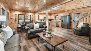 A spacious cabin interior with a wooden interior, a living area with a sofa and coffee table, a kitchen with modern appliances, and a staircase. - NEW BUILD SEPT 2023 4 KINGS 1Q BUNK HOT TUB GAME ROOM THEATER  (Pigeon Forge)
