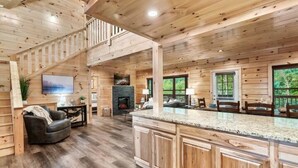 A spacious cabin interior with a large wooden staircase, a fireplace, a kitchen island, and a seating area. - NEW BUILD SEPT 2023 4 KINGS 1Q BUNK HOT TUB GAME ROOM THEATER  (Pigeon Forge)