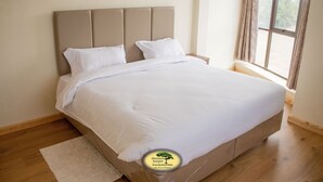 A neatly made bed in a room with a wooden floor, a window with curtains, and a wall-mounted light fixture. - Juniper Green Gardens Resort (Mweiga)