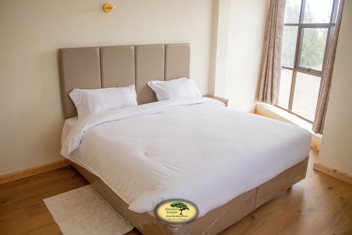 A neatly made bed in a room with a wooden floor, a window with curtains, and a wall-mounted light fixture.