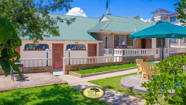 A hotel with a green roof, a covered patio with chairs, and a small garden area. - Juniper Green Gardens Resort (Mweiga)