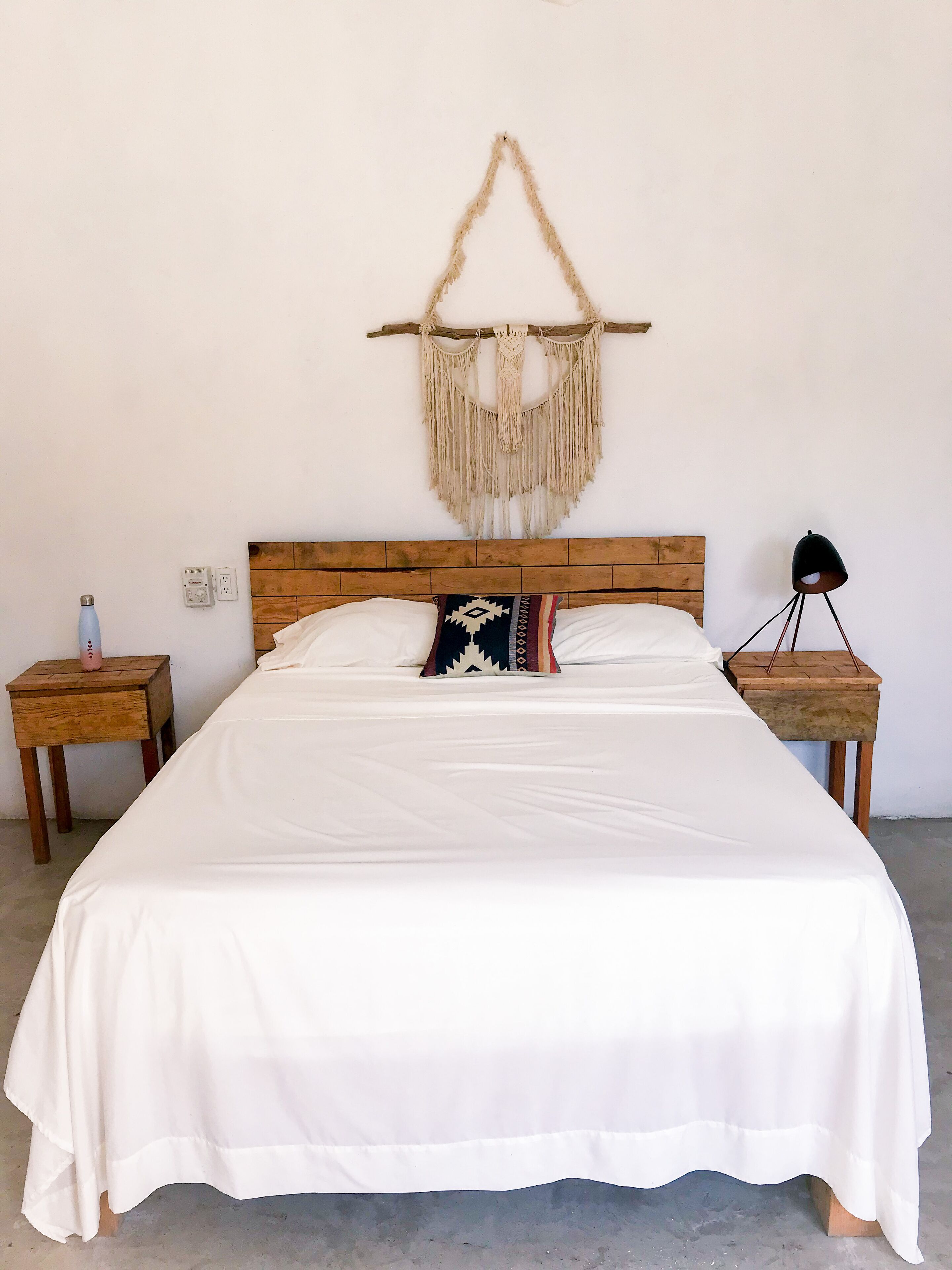 A bed with white bedding, a wooden headboard, a macrame wall hanging, and two bedside tables with lamps.