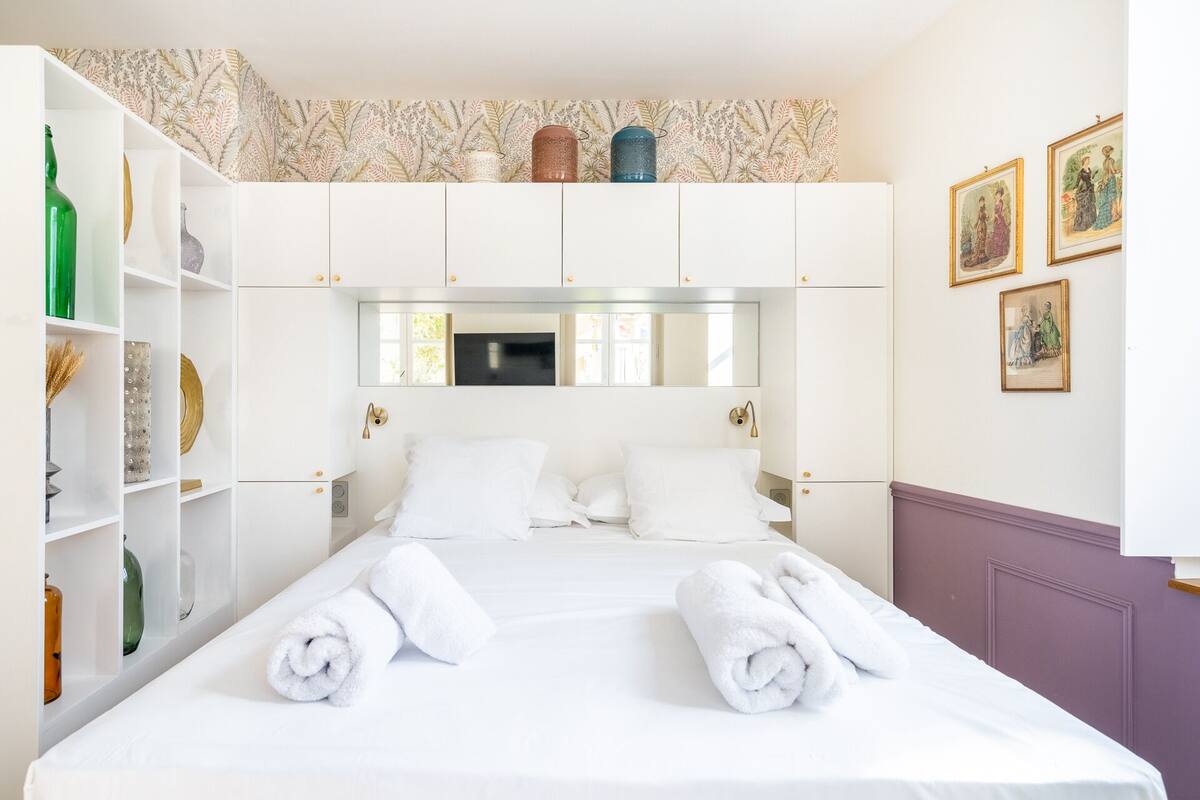 A hotel room with a large bed, white linens, a wardrobe with a mirror, and artwork on the walls.