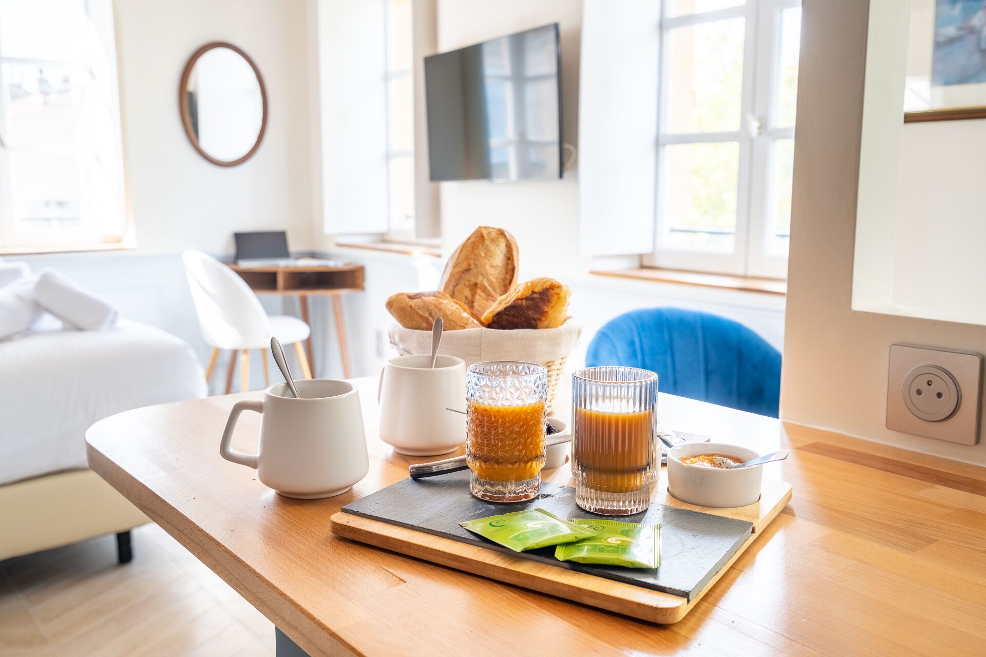 Frühstücksaufstellung mit Brot, einer Tasse, einem Glas Orangensaft, einem Glas Kaffee und einer Schüssel Müsli auf einem Holztisch in einem hellen Raum mit einer Couch und einem Fernseher im Hintergrund.