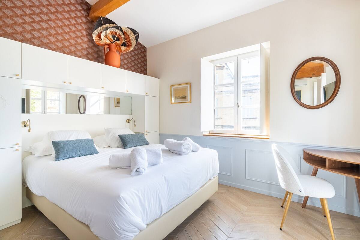 A bedroom with a bed, a chair, a desk, a mirror, and a window.