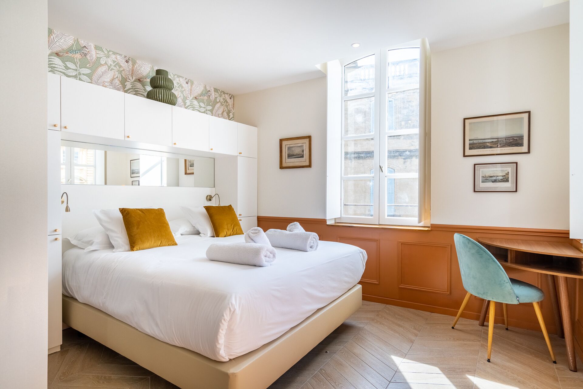 Un dormitorio con una cama, un escritorio y una silla. Hay dos ventanas y un espejo en la pared.