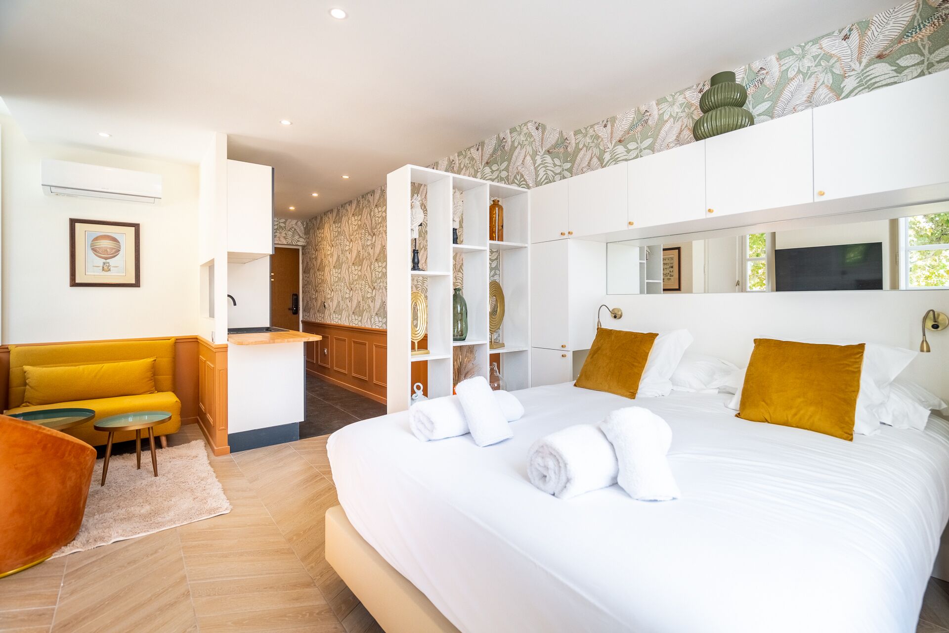 A modern hotel room with a large bed, a small seating area, a kitchenette, and a bathroom.