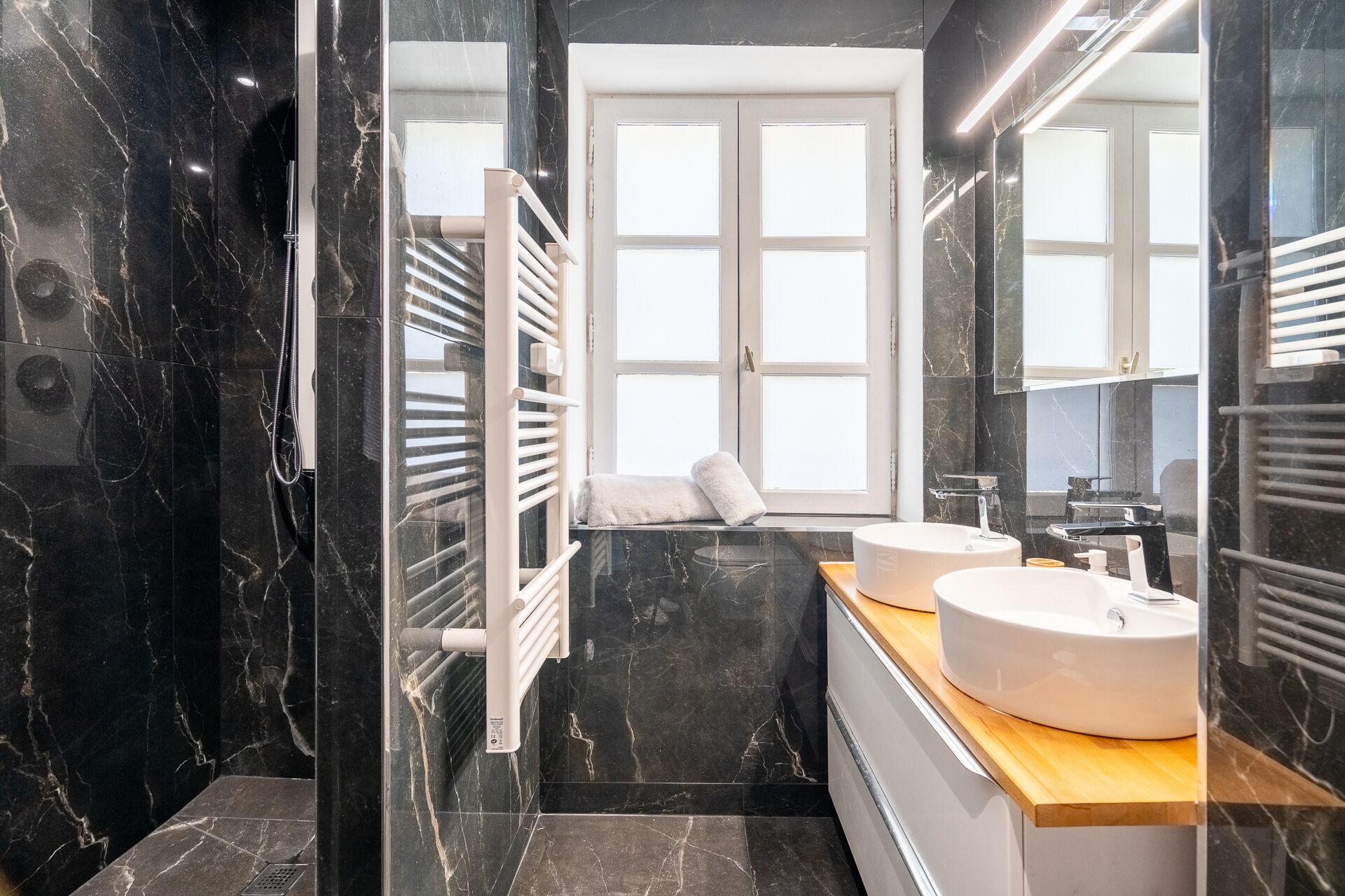 Une salle de bain moderne avec des murs en marbre noir, deux vasques blanches, un meuble-lavabo en bois et un grand miroir.