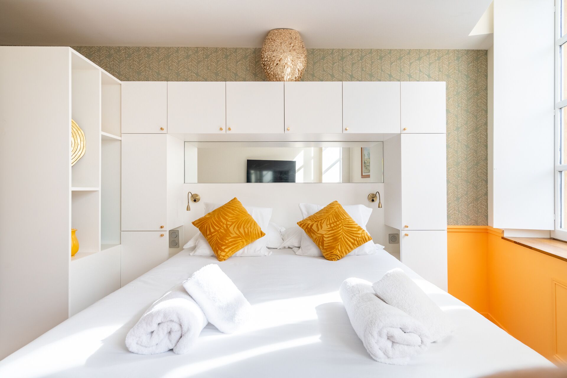 A hotel room with a large bed, white bedding, two yellow pillows, a mirror above the bed, and a window with orange walls.