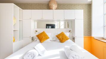 A hotel room with a large bed, white bedding, two yellow pillows, a mirror above the bed, and a window with orange walls.