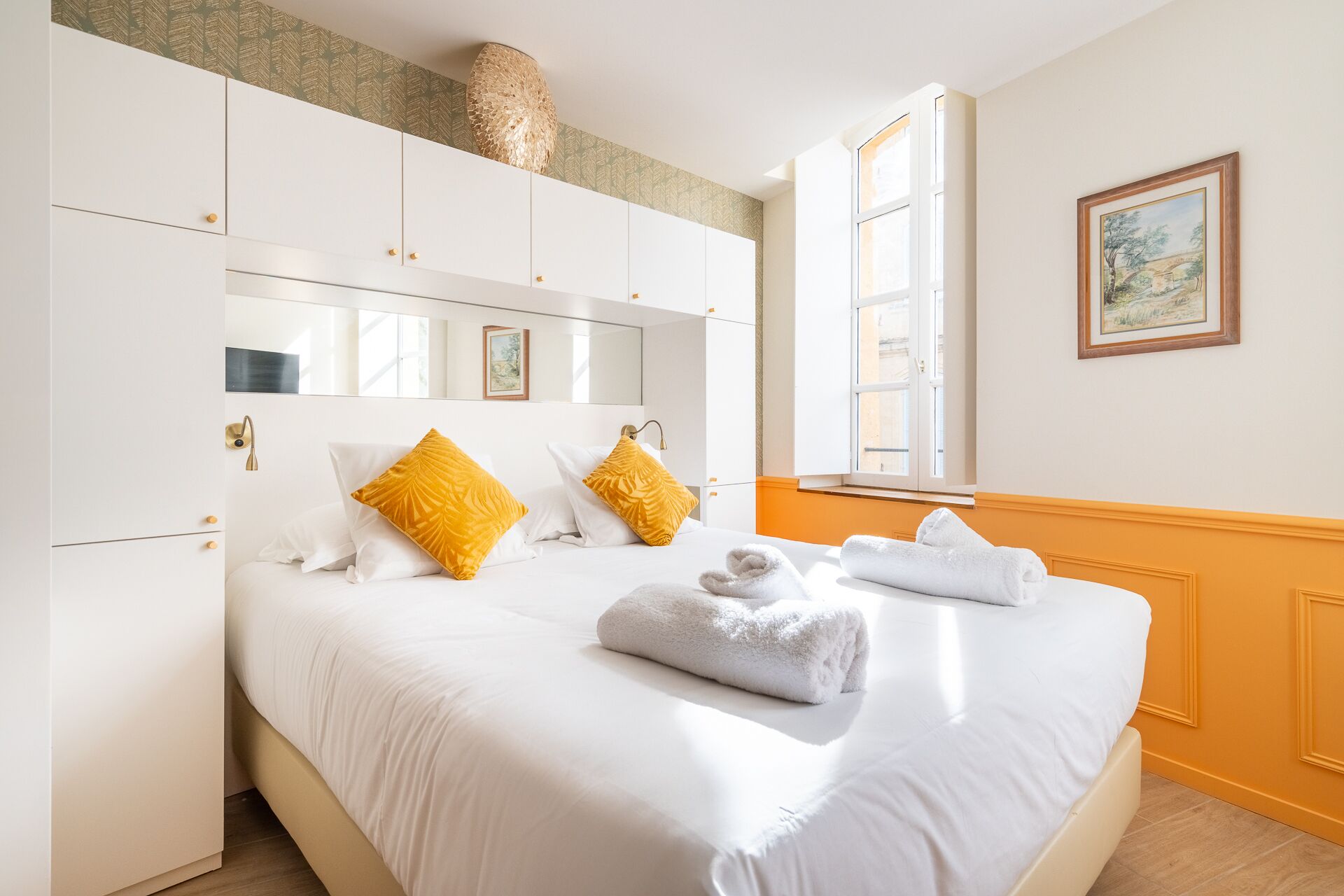 A hotel room with a large bed, white bedding, two yellow pillows, a large window, and a painting on the wall.