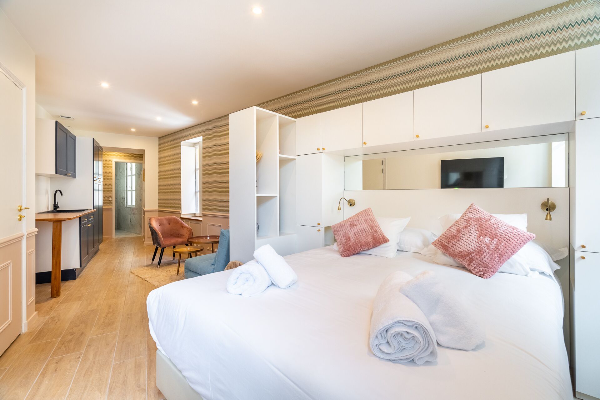 Una moderna camera d'albergo con un letto grande, un'area salotto con una poltrona, una piccola scrivania e un bagno visibile attraverso una porta aperta.