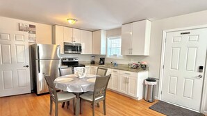 A compact kitchen with white cabinets, a stainless steel refrigerator, a microwave, and a stove. There is a dining table with four chairs and a small trash can near the door.