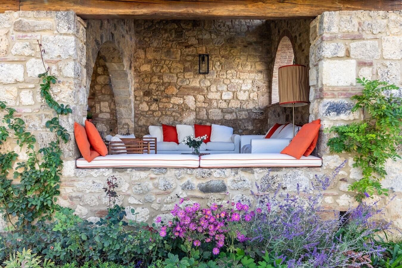 A stone-walled outdoor seating area with cushions and a table, surrounded by greenery and flowers.