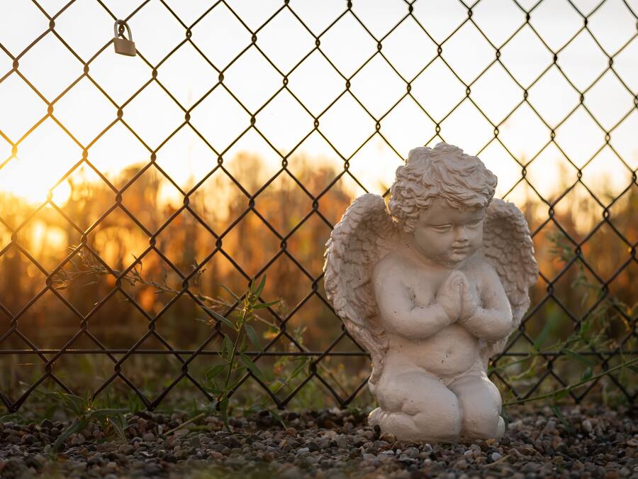 Une statue d'un ange, placée derrière une clôture en treillis métallique, avec un coucher de soleil flou en arrière-plan.