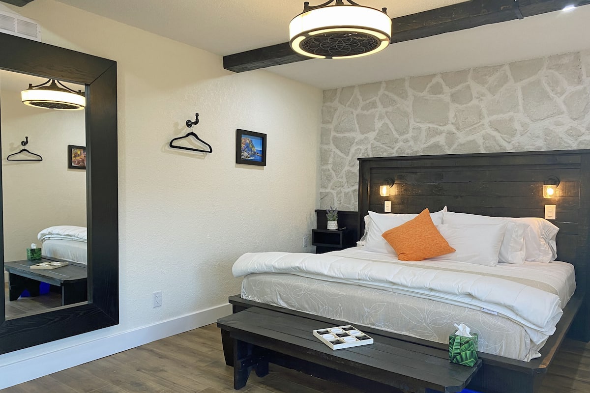 Una habitación de hotel con cama, una banca, un espejo, una mesita de noche y un aplique de pared.
