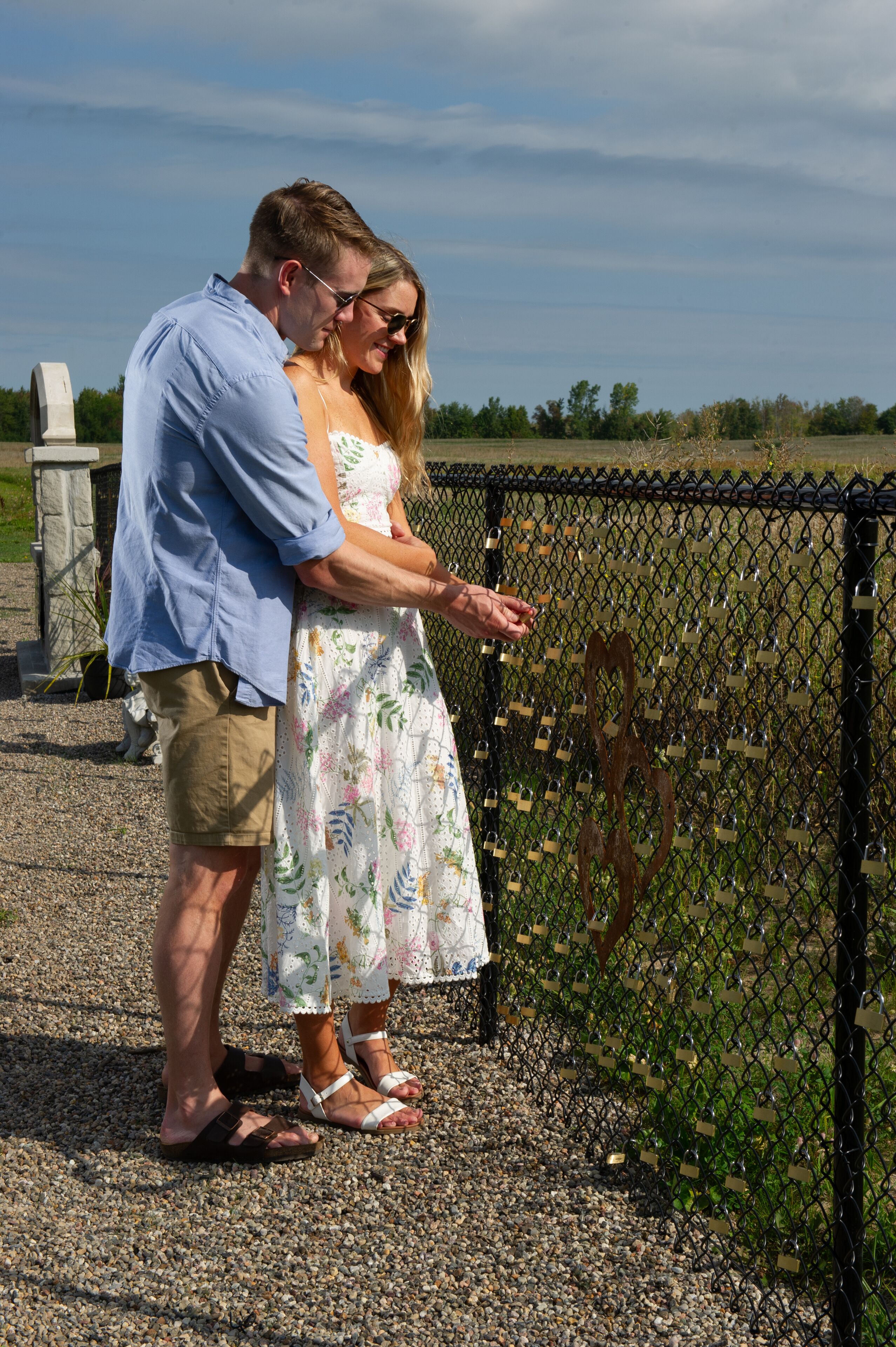 A couple standing by a chain-link fence with padlocks, a gravel path, and a clear sky in the background.