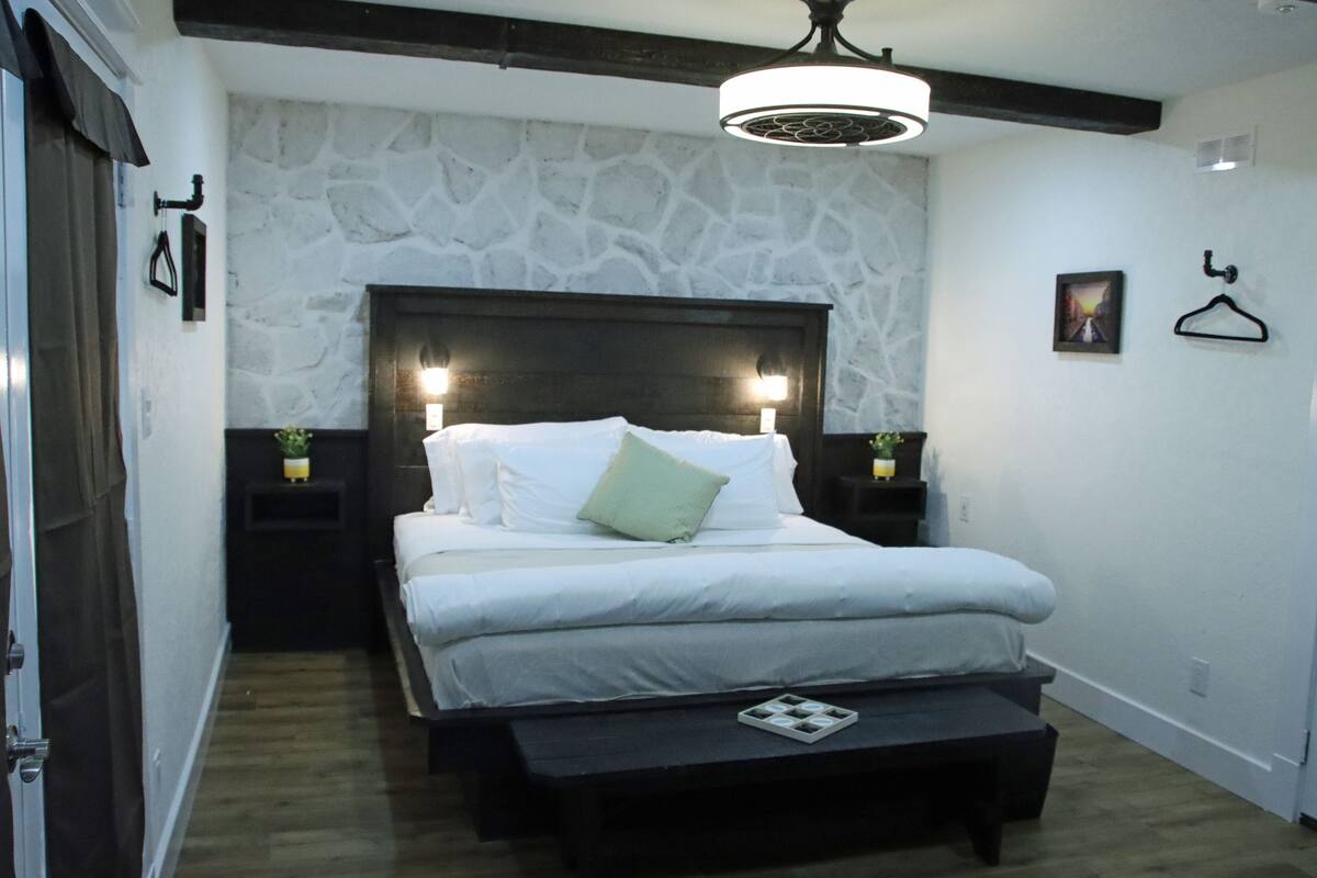 Un dormitorio con pared de piedra, cabecera de madera, mesita de noche con lámpara y perchero.