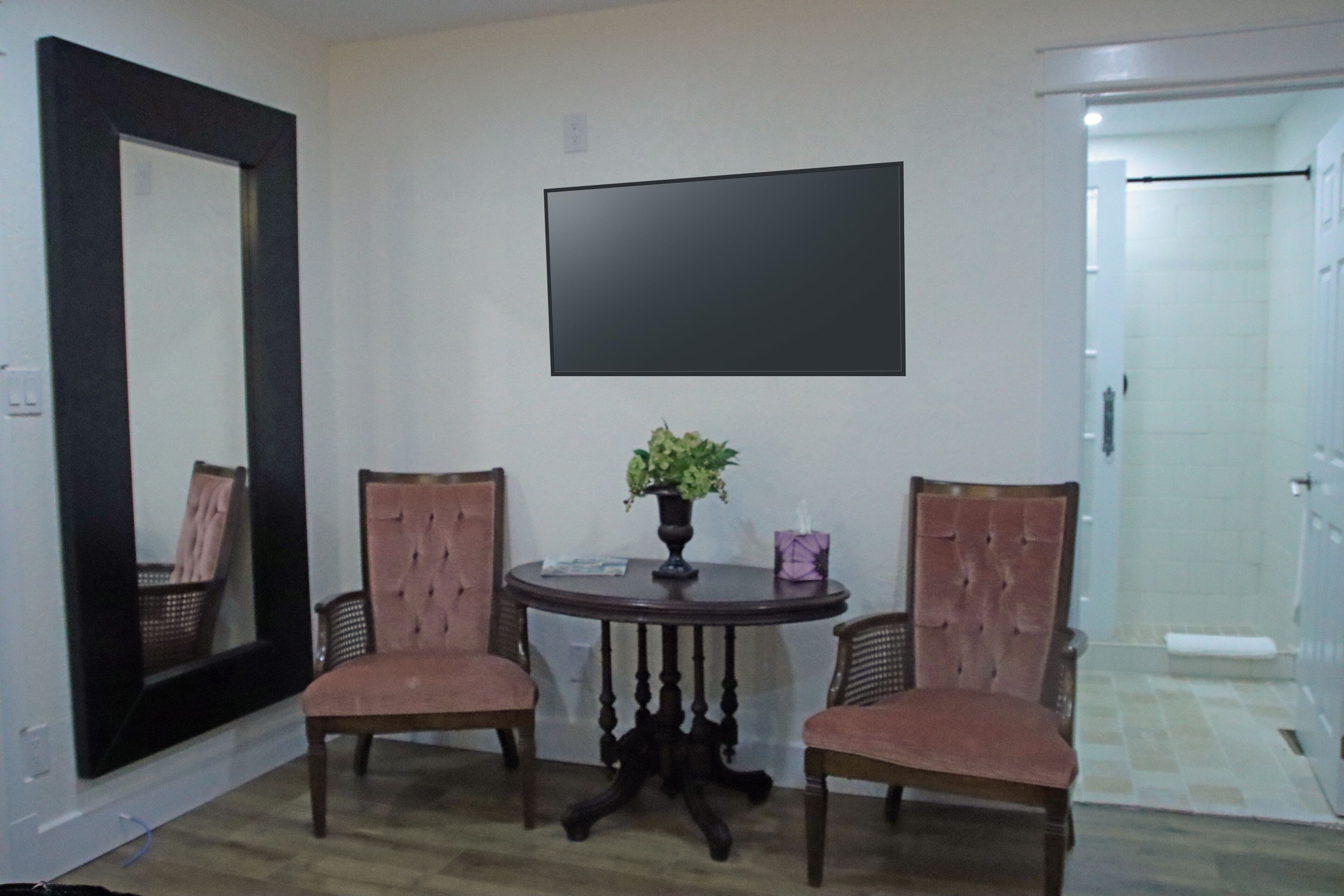 A room with a round table, two chairs, a potted plant, a flat-screen TV, and a door leading to a bathroom.