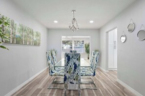 A dining area with a glass-top table, chairs with floral upholstery, a chandelier, and a view through a window.