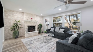 A living room with a fireplace, a ceiling fan, a sofa, and a large window with blinds. - Spacious House w/ Patio & Fenced Yard (Lakeland)
