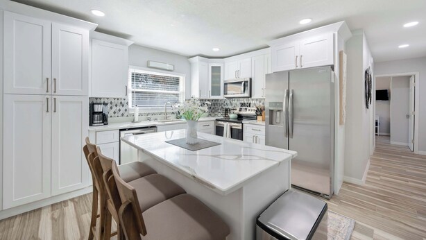 A modern kitchen with white cabinets, a central island, stainless steel appliances, and a window with blinds. - Spacious House w/ Patio & Fenced Yard (Lakeland)