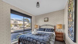 Une chambre avec une grande fenêtre, un lit recouvert d’une couvre-lit à motifs, deux lampes de chevet, une table de chevet en osier et une œuvre d’art murale.
