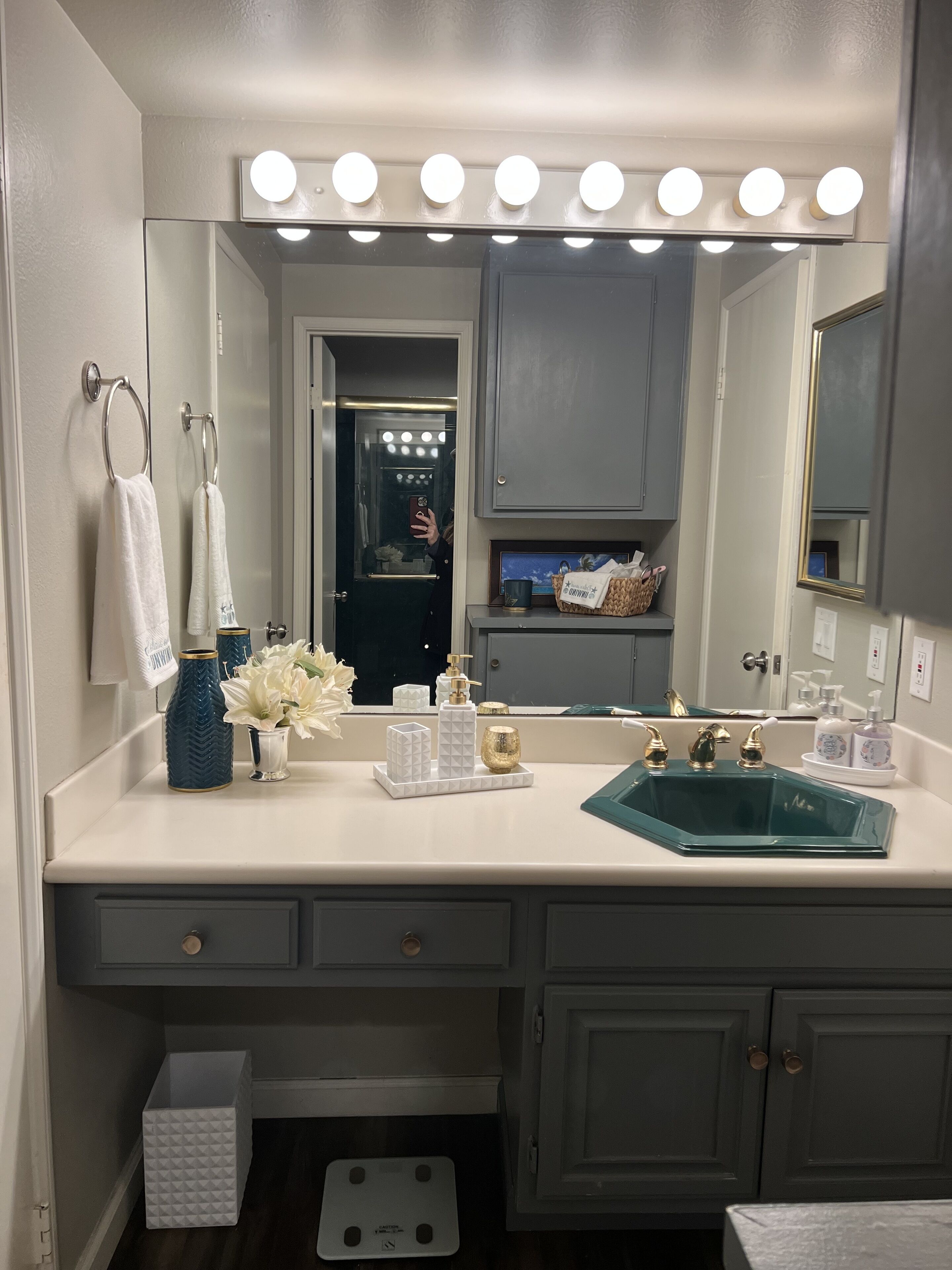 A bathroom with a vanity, a sink, a mirror with lights, a towel rack, and a trash bin.