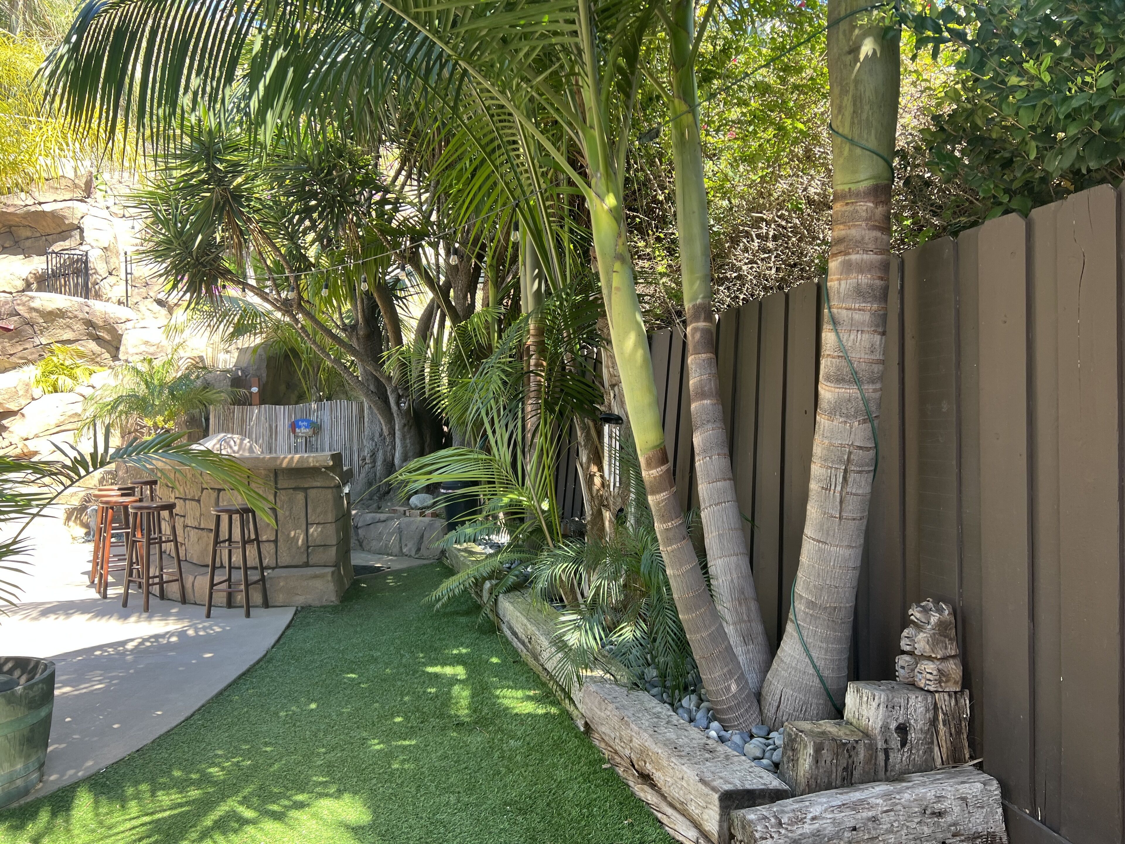 A backyard with artificial grass, a wooden bench, a stone wall, and a variety of tropical plants.