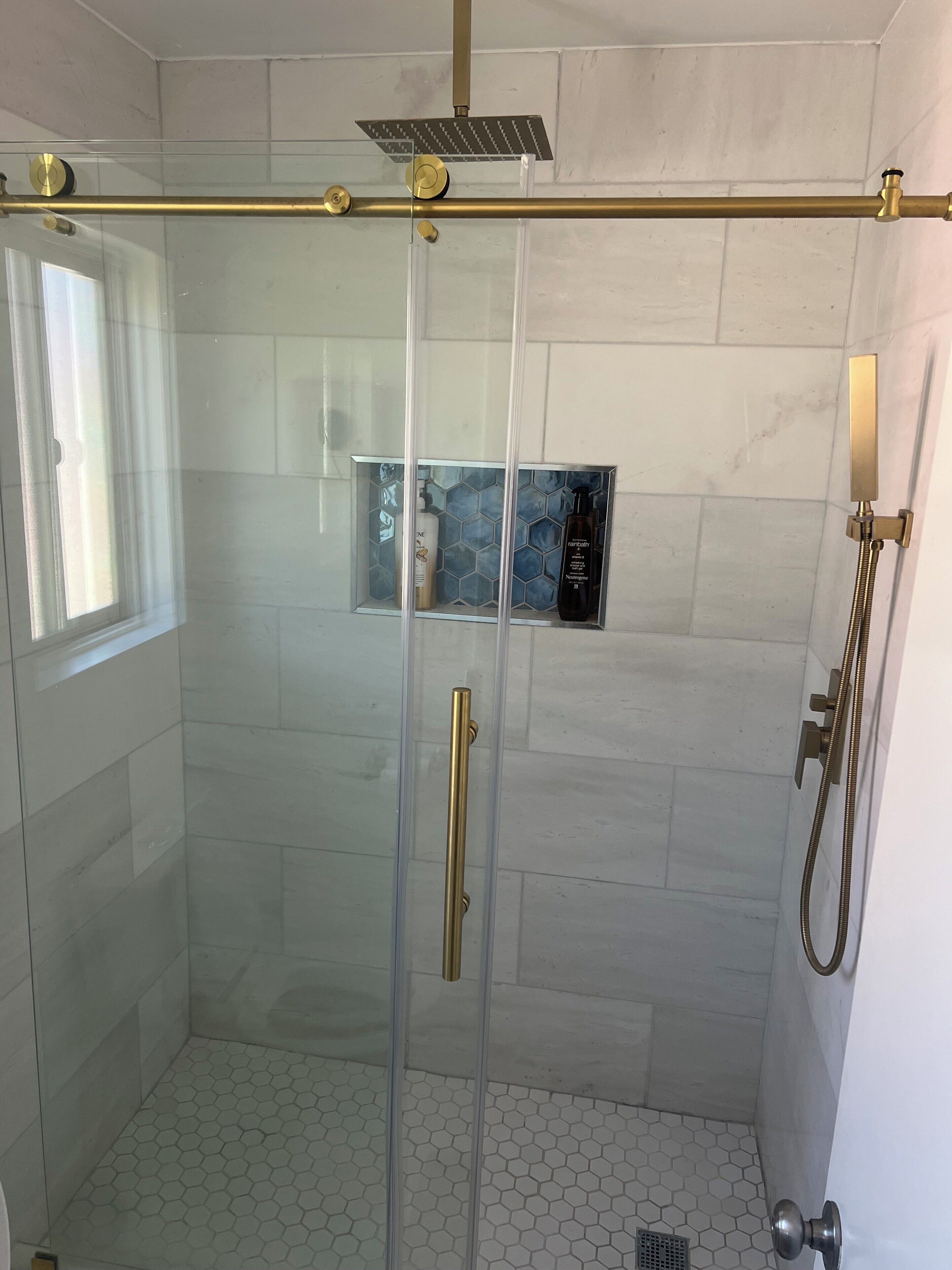 Ducha con cerramiento de vidrio, grifo dorado, ducha de lluvia y repisa con artículos de aseo.
