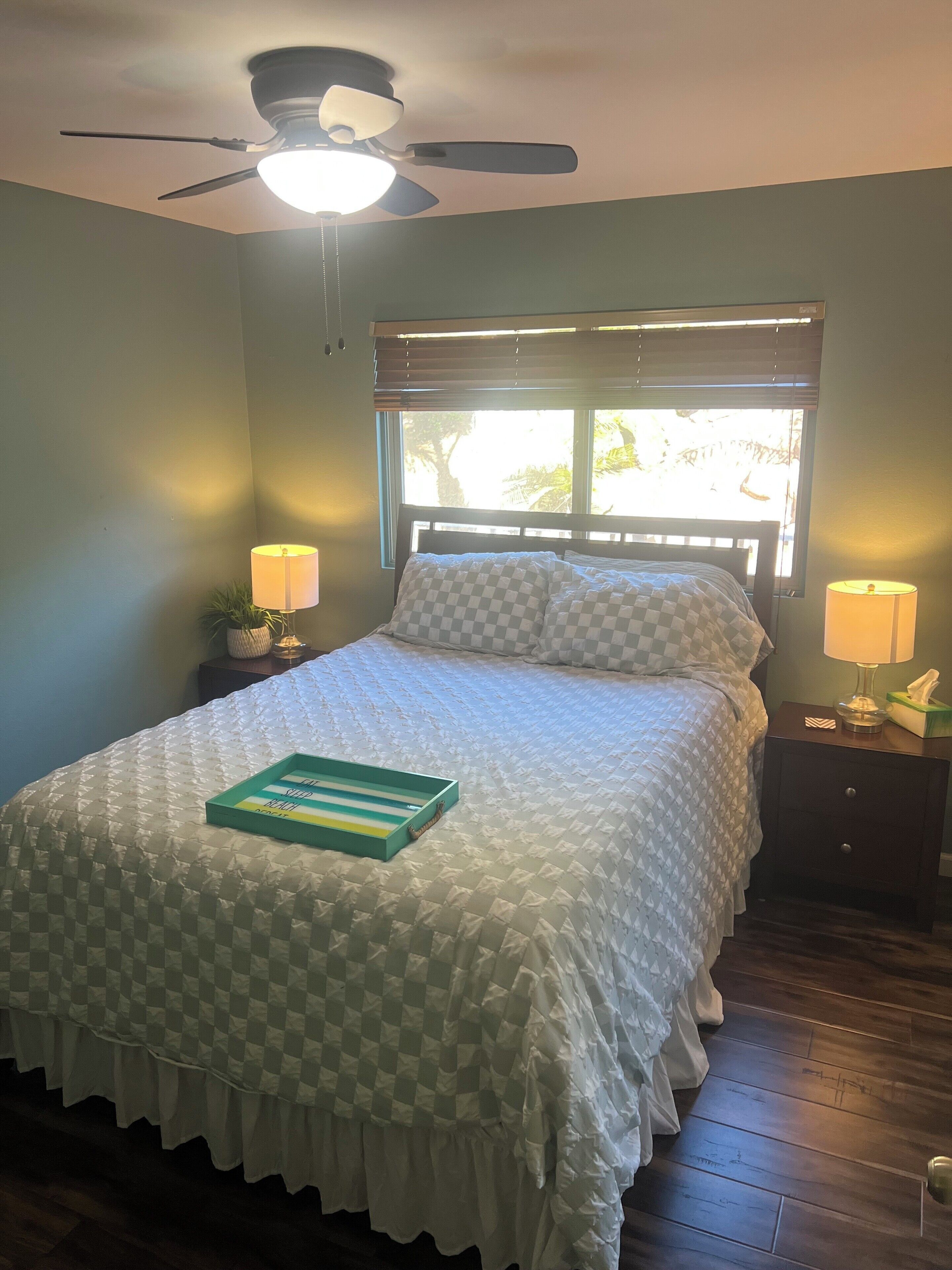 Un dormitorio con cama, ventilador de techo, ventana con persianas y lámparas de noche.