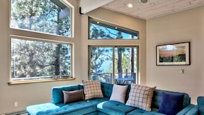 Un salon moderne avec un canapé d’angle turquoise, de grandes fenêtres donnant sur la verdure et un tableau encadré accroché au mur.