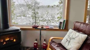 Une pièce chaleureuse avec un poêle à bois, un canapé en cuir agrémenté d’un coussin décoratif, une fenêtre donnant sur des arbres couverts de neige et une petite table avec des livres et une théière.