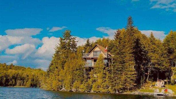 Une cabane entourée d’une forêt dense, avec un ciel dégagé et une étendue d’eau au premier plan.