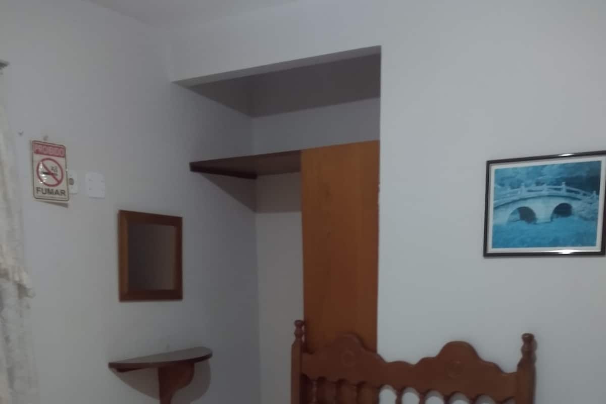 Un dormitorio con cama de madera, ropa de cama blanca, una mesita de noche pequeña, un estante en la pared, un cuadro enmarcado y un cartel de 'No Fumar'.