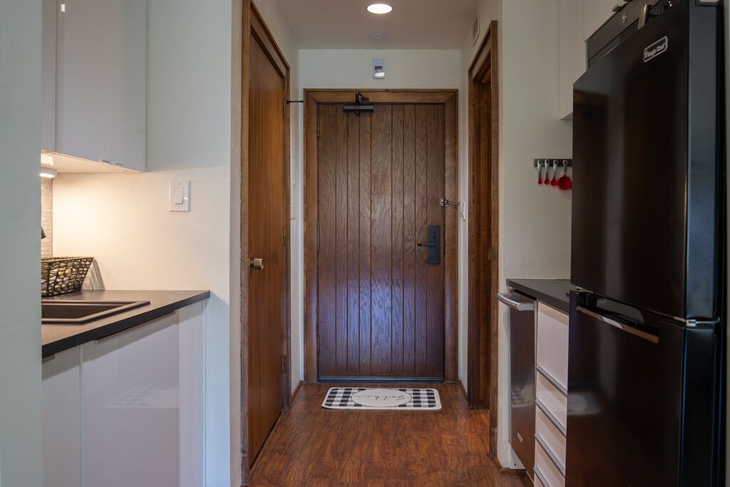 A narrow hallway with a wooden door at the end, a black refrigerator on the right, and a small kitchen area with cabinets and a countertop on the left.