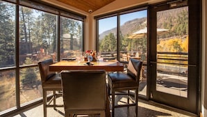 Un comedor con mesa y sillas de madera, ventanas amplias con vistas a árboles y colinas, y un letrero de salida.