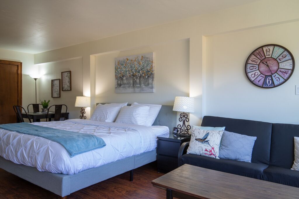 A bedroom with a bed, a sofa, a coffee table, a wall clock, a painting, and a lamp.