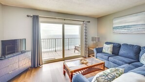 A living room with a television, sofa, coffee table, and a balcony with a view of the sea. - Steps to Shore: PCB Condo w/ Pool Access + Views! (Panama City Beach)