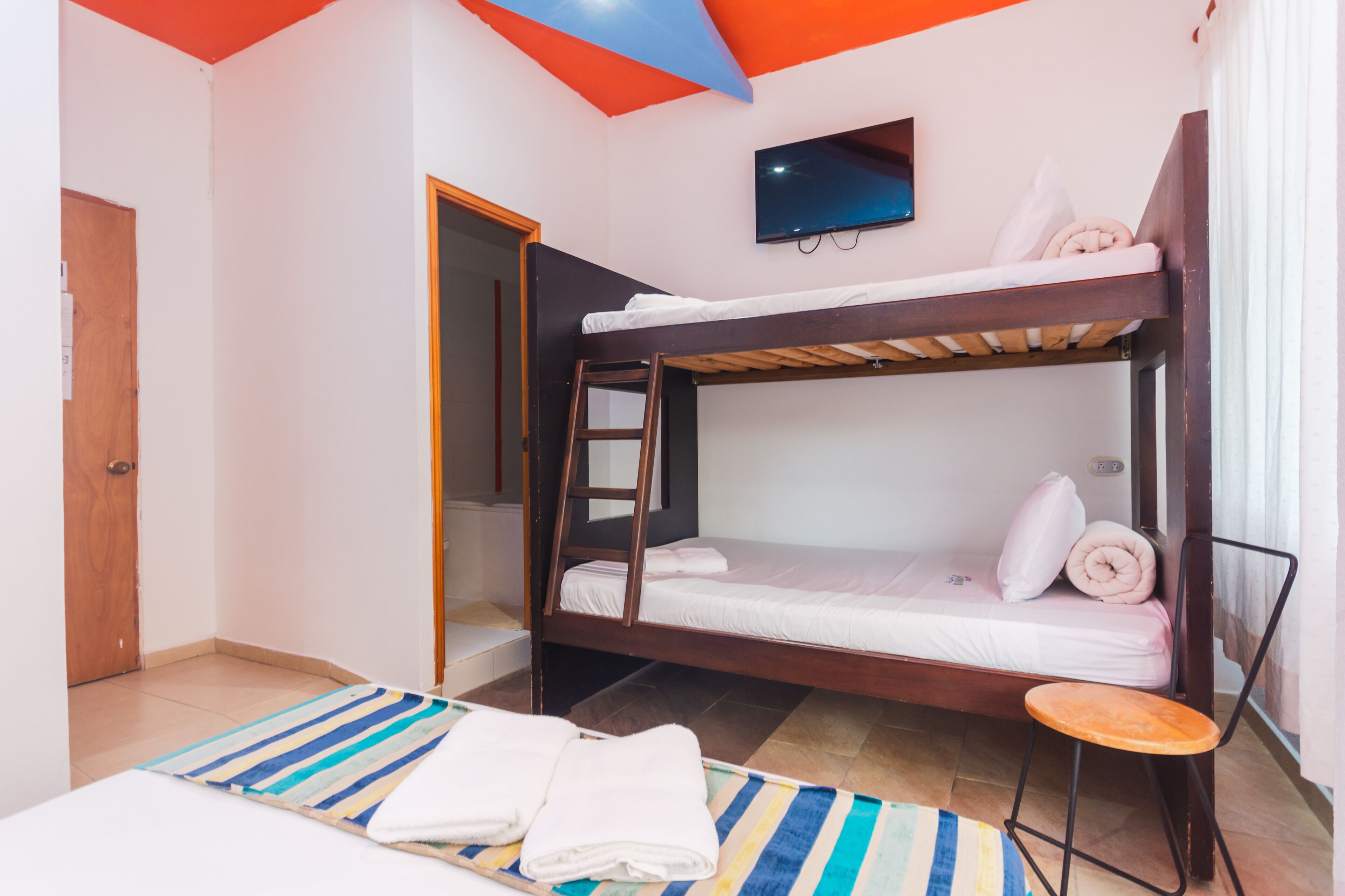 Ein Schlafraum mit Etagenbetten, einem Fernseher über dem oberen Bett, einer Holztür und einem Streifenmuster-Teppich auf dem Boden.
