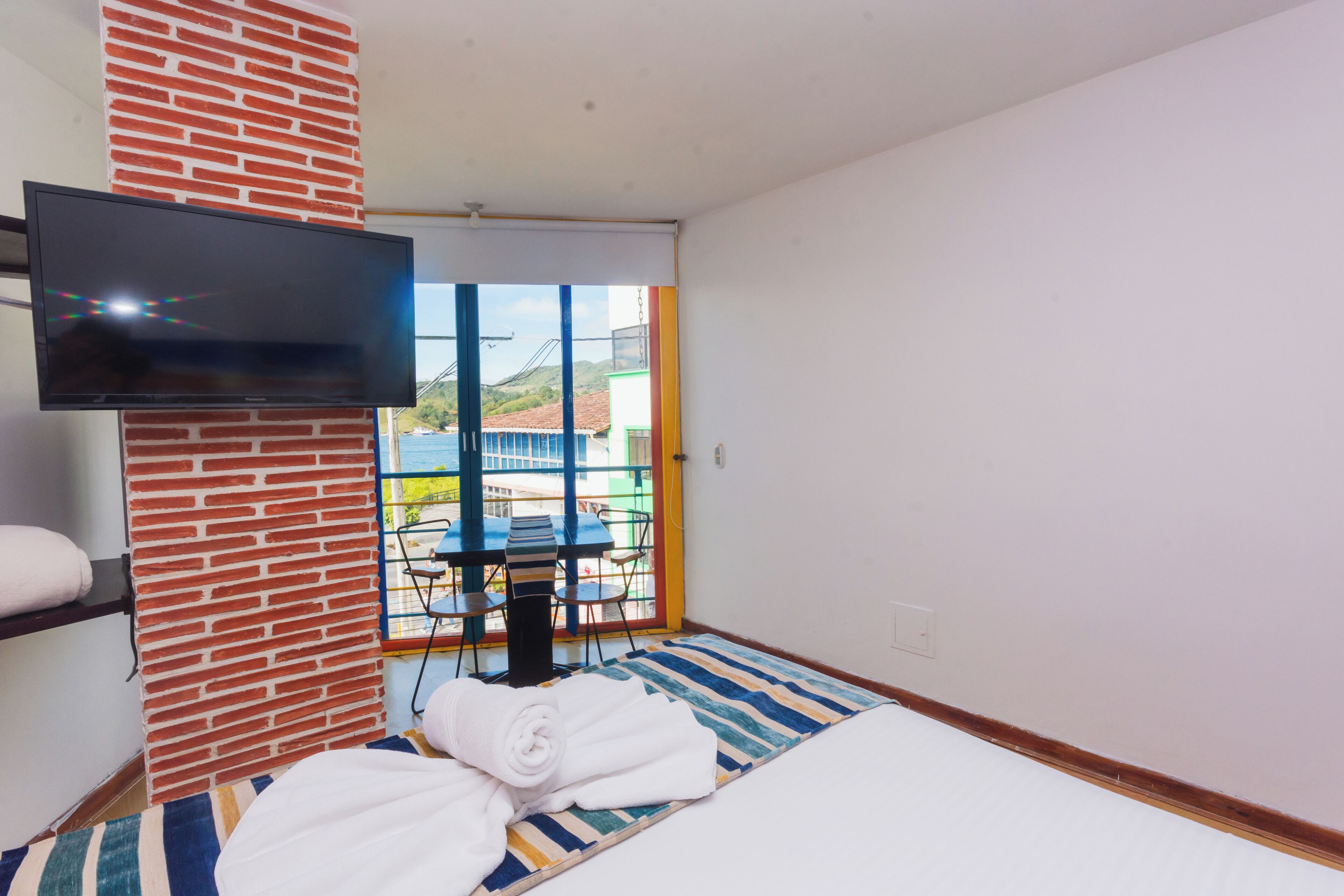 Camera d'albergo con parete decorata in mattoni, televisione a schermo piatto sopra il letto, tappeto a righe e vista sull'esterno attraverso un'ampia finestra.