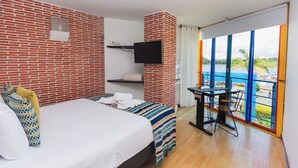 A hotel room with a brick wall, a bed with a striped blanket, a TV mounted on the wall, a desk with a chair, and a large window with a view of the sea. - Hotel Guatatur (Guatapé)