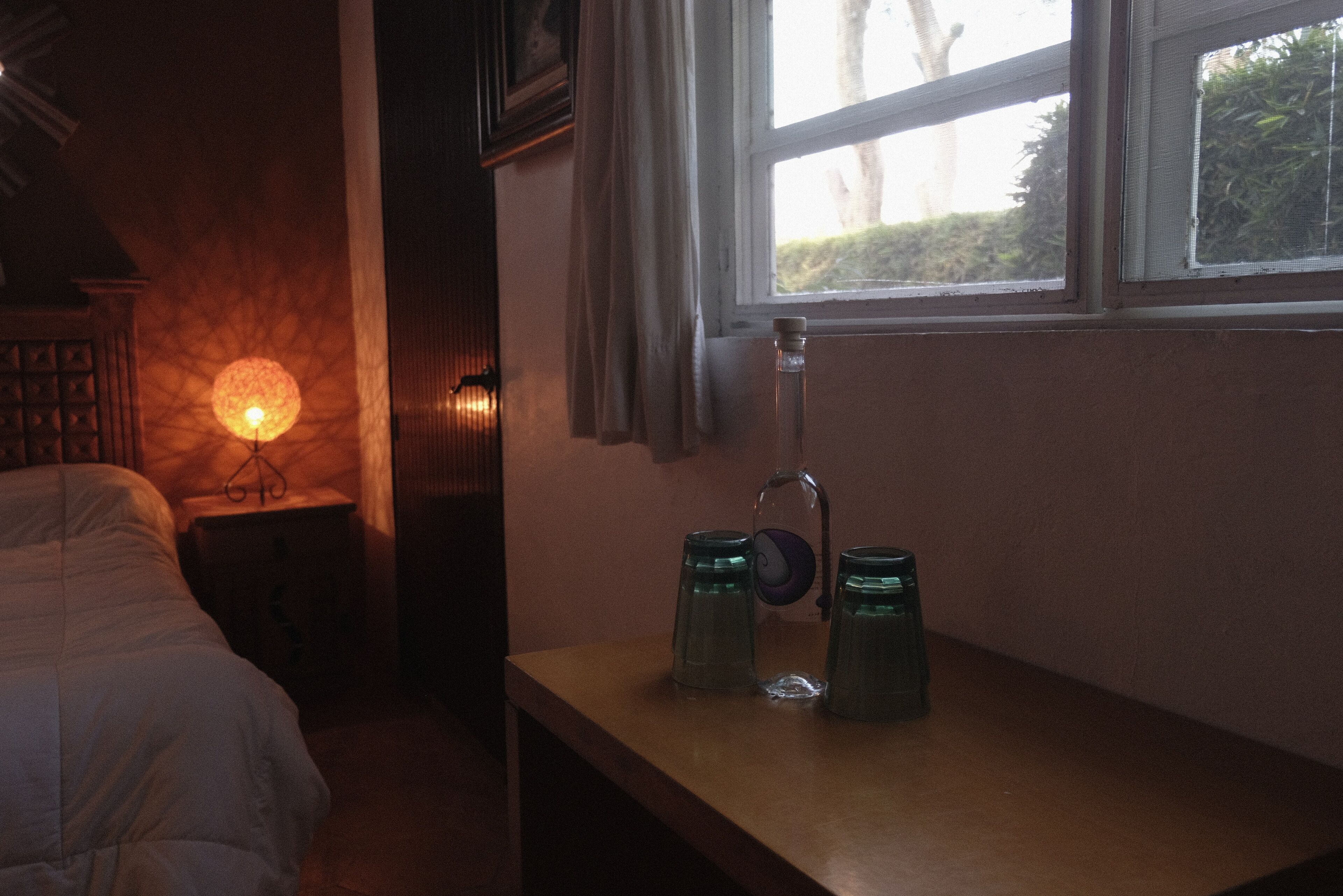 Ein Schlafzimmer mit einem Bett, einem Nachttisch mit Lampe und einem Fenster mit Blick auf Grünflächen.