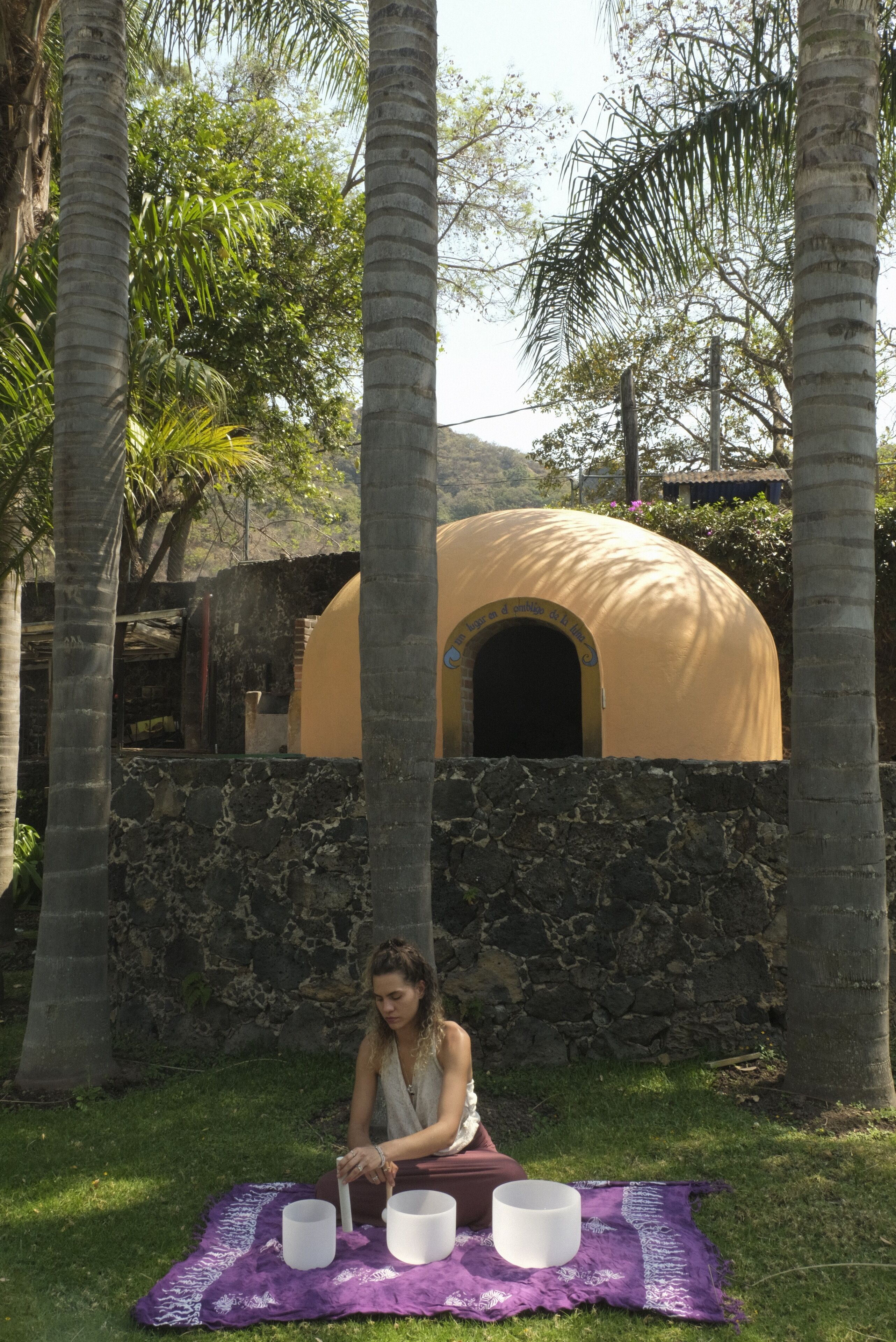 A person sitting on a blanket with three white cups in front of a stone wall and a yellow dome structure in the background.