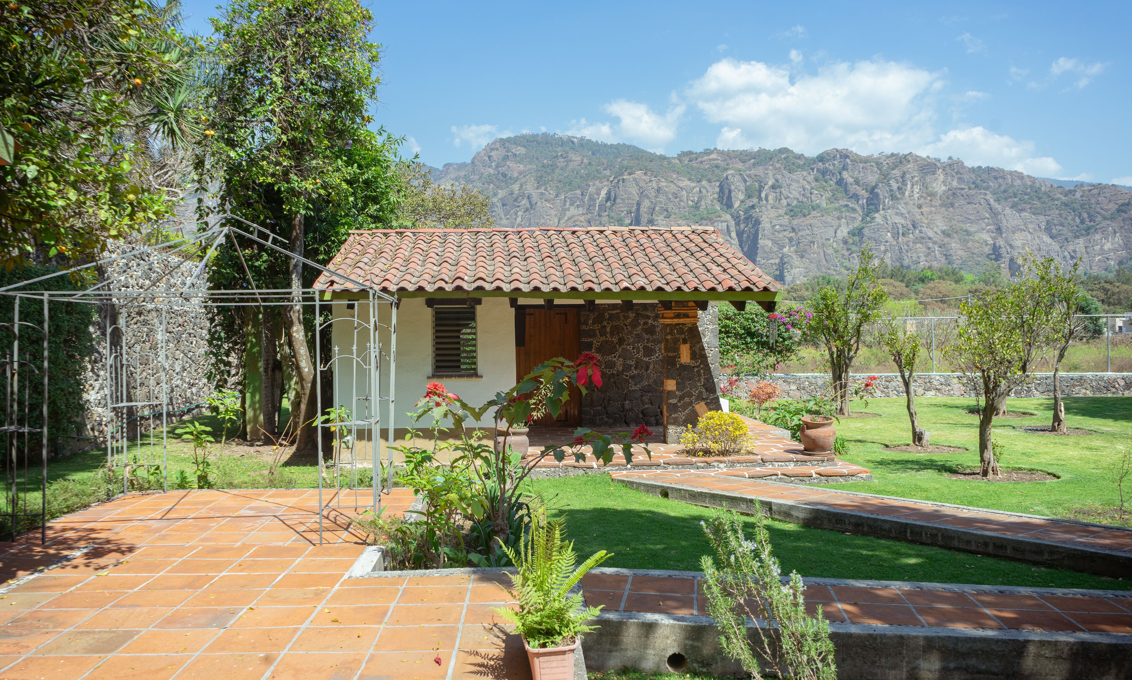 A small house with a tiled roof, surrounded by a garden with various plants and a view of mountains in the background.