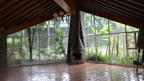 Amplia habitación con una gran chimenea de piedra, vigas de madera y ventanas amplias que dan a vistas de vegetación.