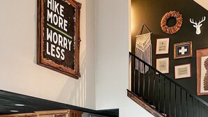 A living room with a brown leather sofa, a kitchen with wooden cabinets, and a staircase with a decorative wreath and a deer head. - Pine Lane is your cozy, pet-friendly escape -w/ hot tub, fire pit, & peaceful vibes near Mohican (Butler)